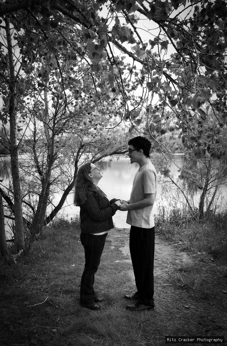 Black-and-white portrait of two people holding hands beneath trees beside water.