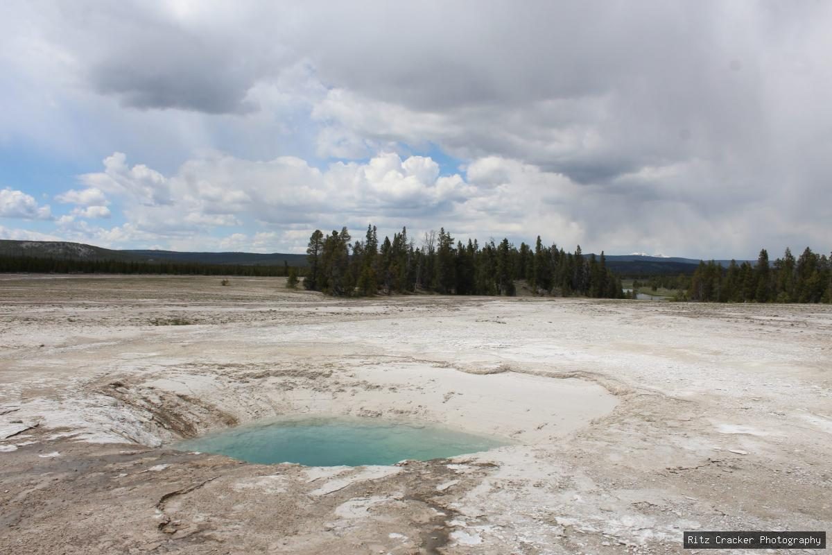 Small turquoise hot spring in a pale mineral basin, with forest and broad clouds in the distance.